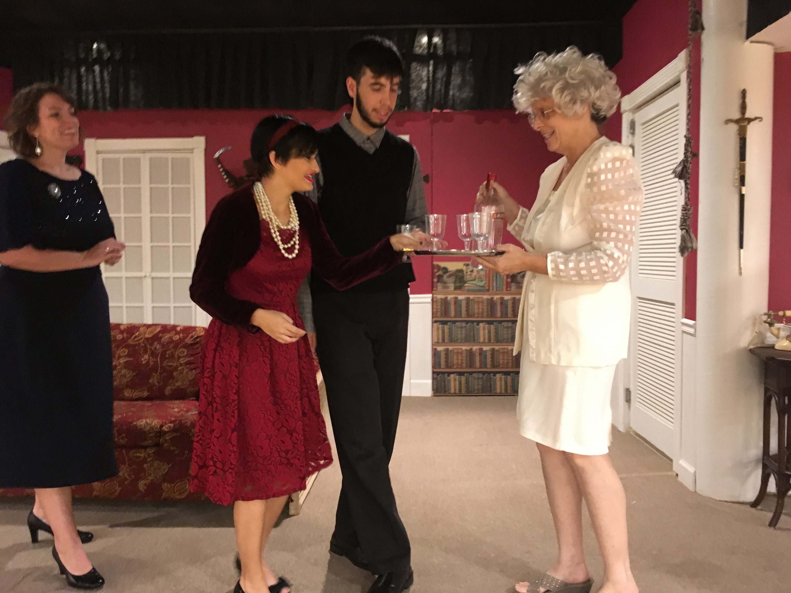A female actor holds a tray with wine glasses and is offering them to a male actor and two female actors