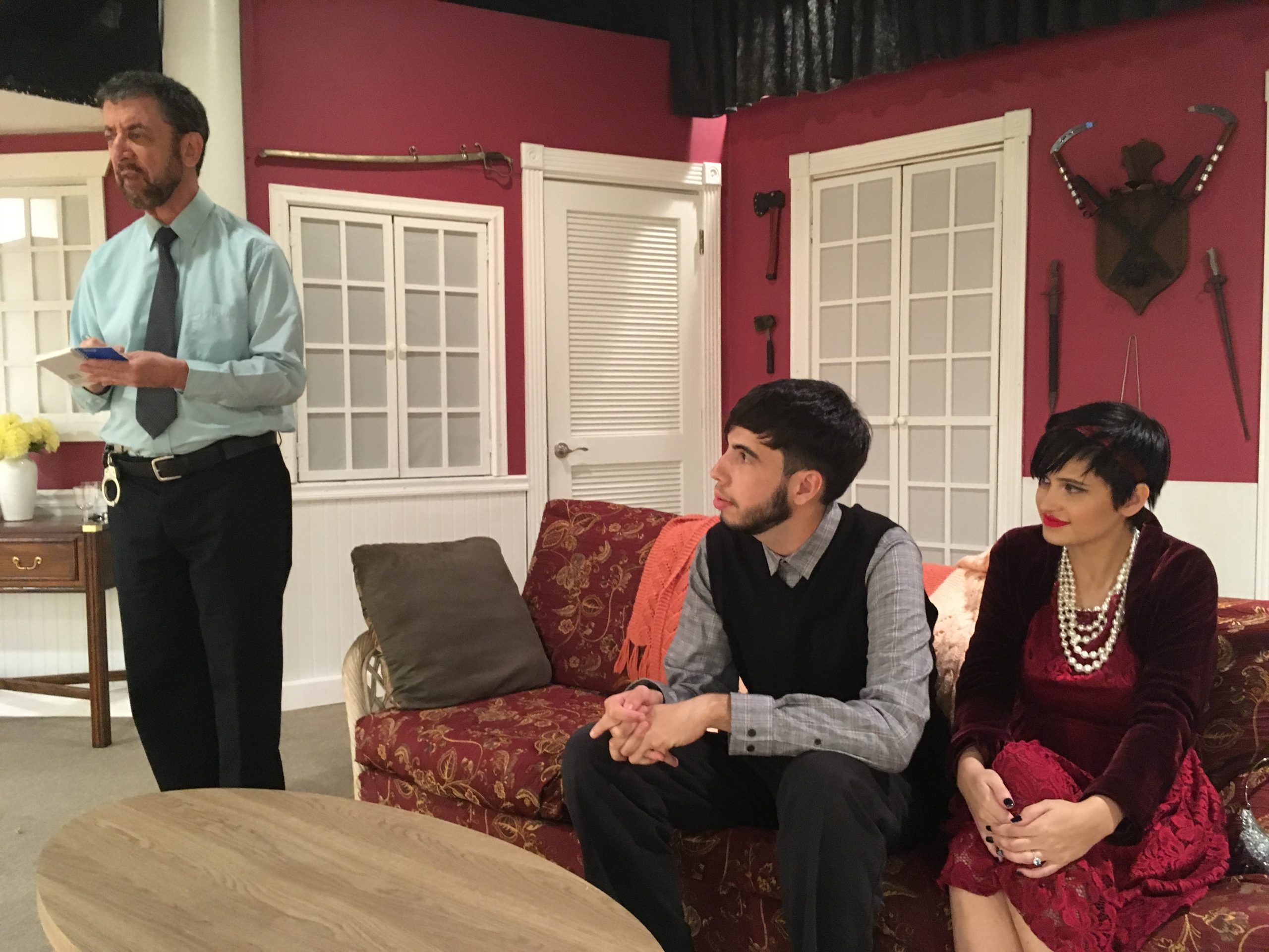A male actor stands, holding a notebook while a male and female actor sit on the couch watching him.