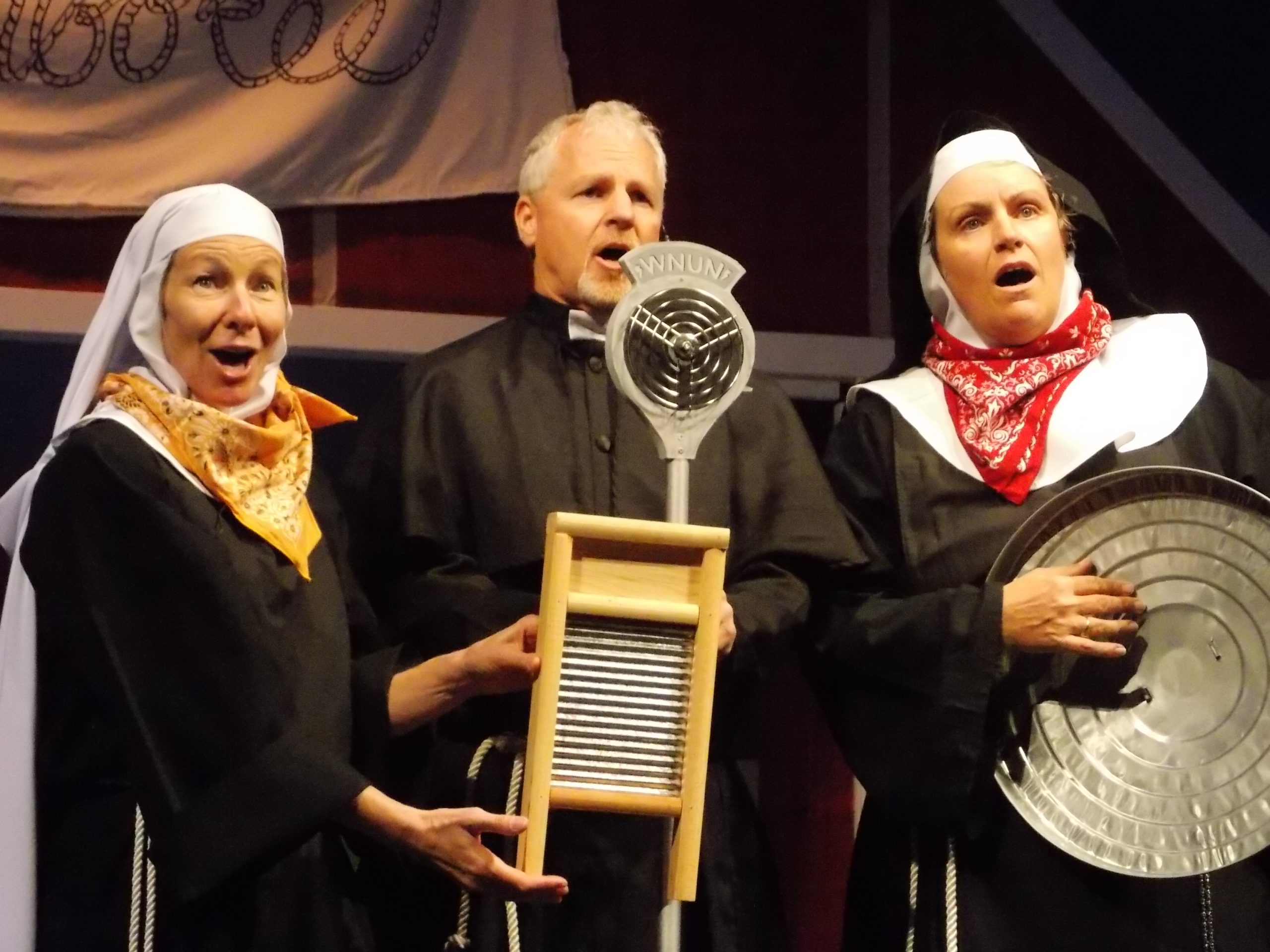 Two female actors dressed in nuns habits on either side of a male actor in a priest uniform holding a washboard