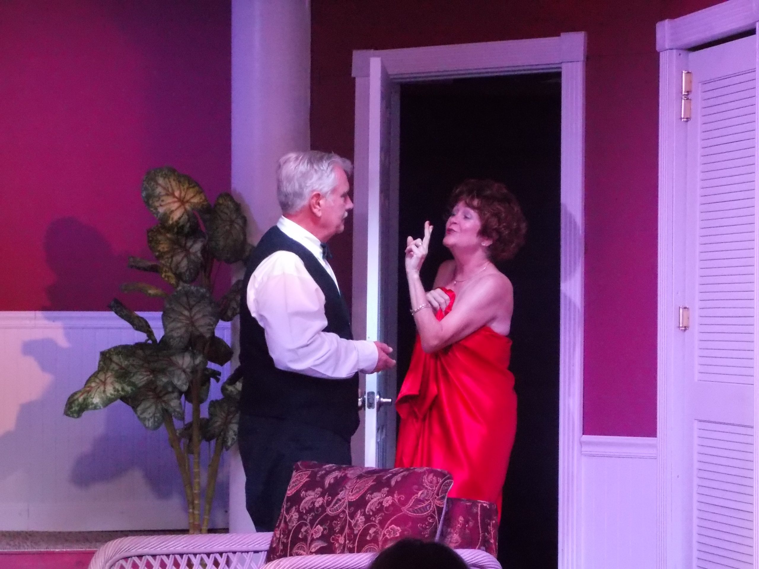 A male actor dressed in a white shirt and black tuxedo opens a door and a female actor in a red dress walks in