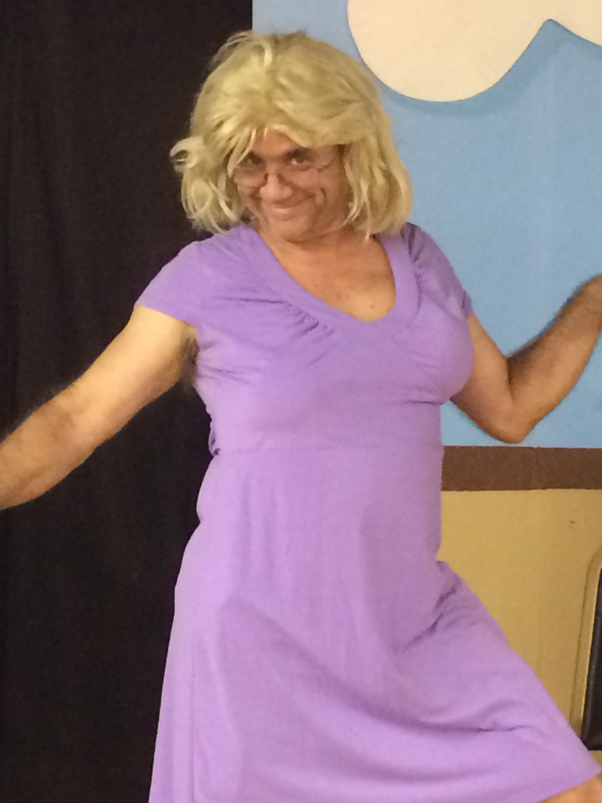 A male actor dressed as a woman in a purple dress and blonde wig