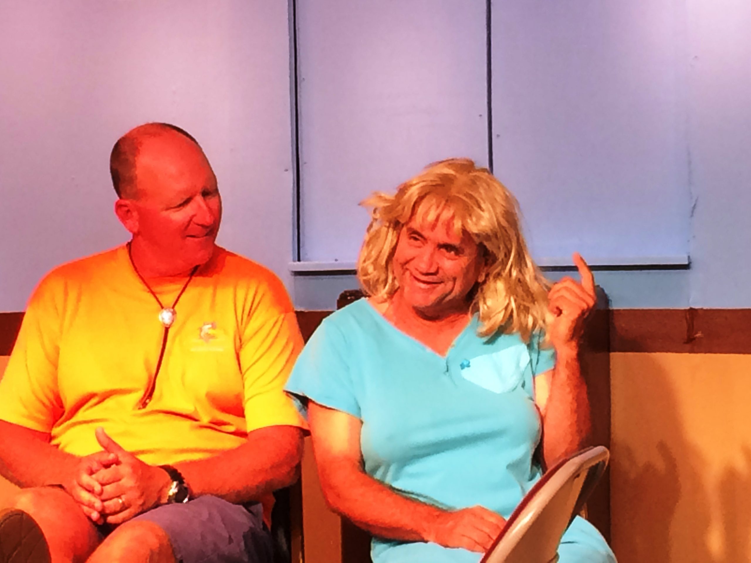 A male actor wearing a yellow shirt sits on a chair and talks with a male actor dressed as a female actor wearing a blue dress and blonde wig