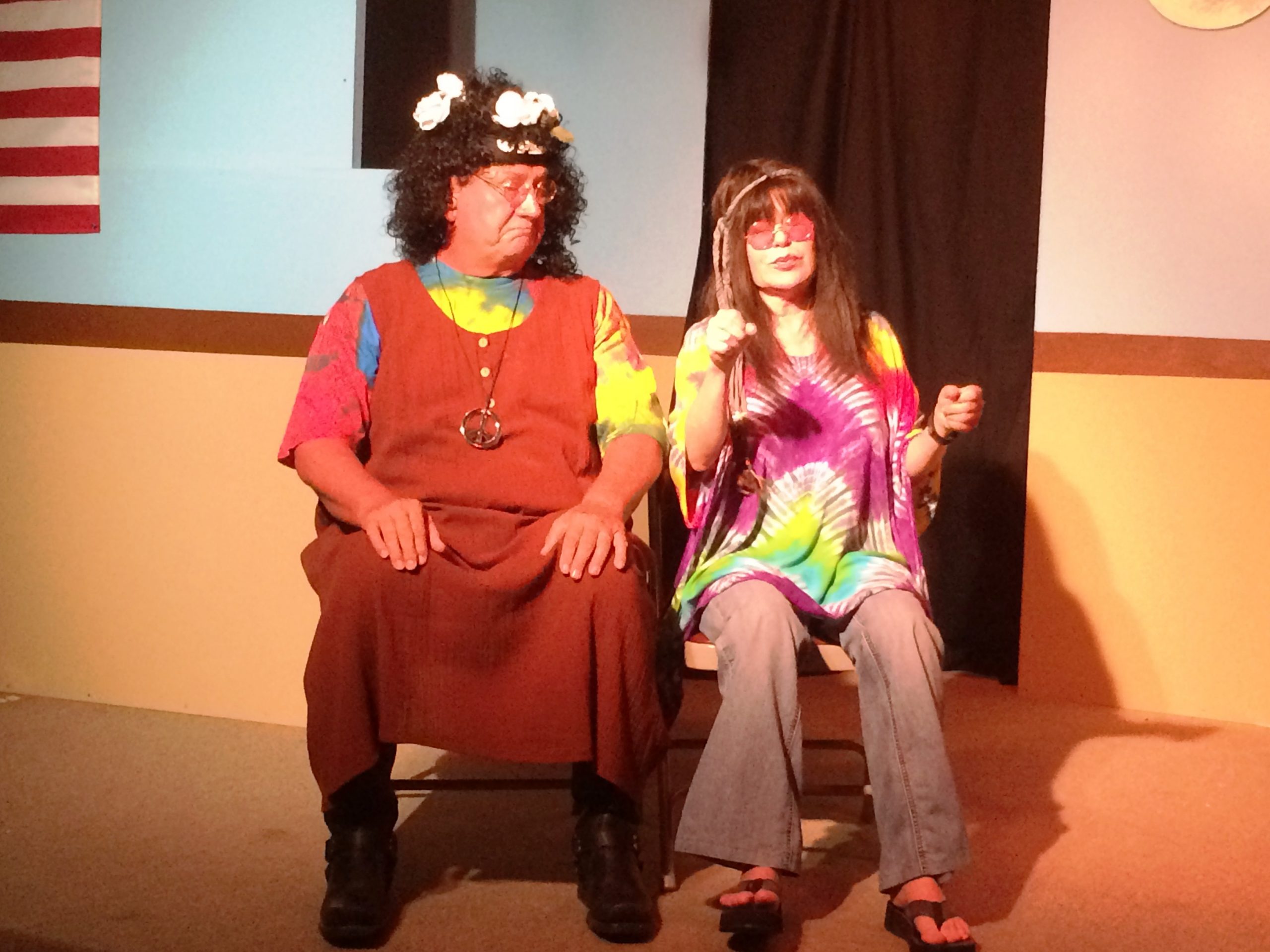 A male actor wearing a funny hat sits next to a female actor wearing a tie-dyed shirt, wide legged pants, and sunglasses