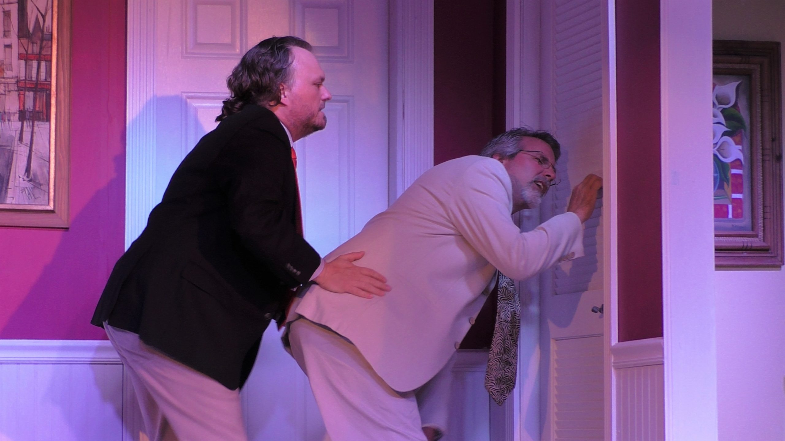 Two male actors stand outside a door. One is leaned over to try to hear what is occurring on the other side of the door.