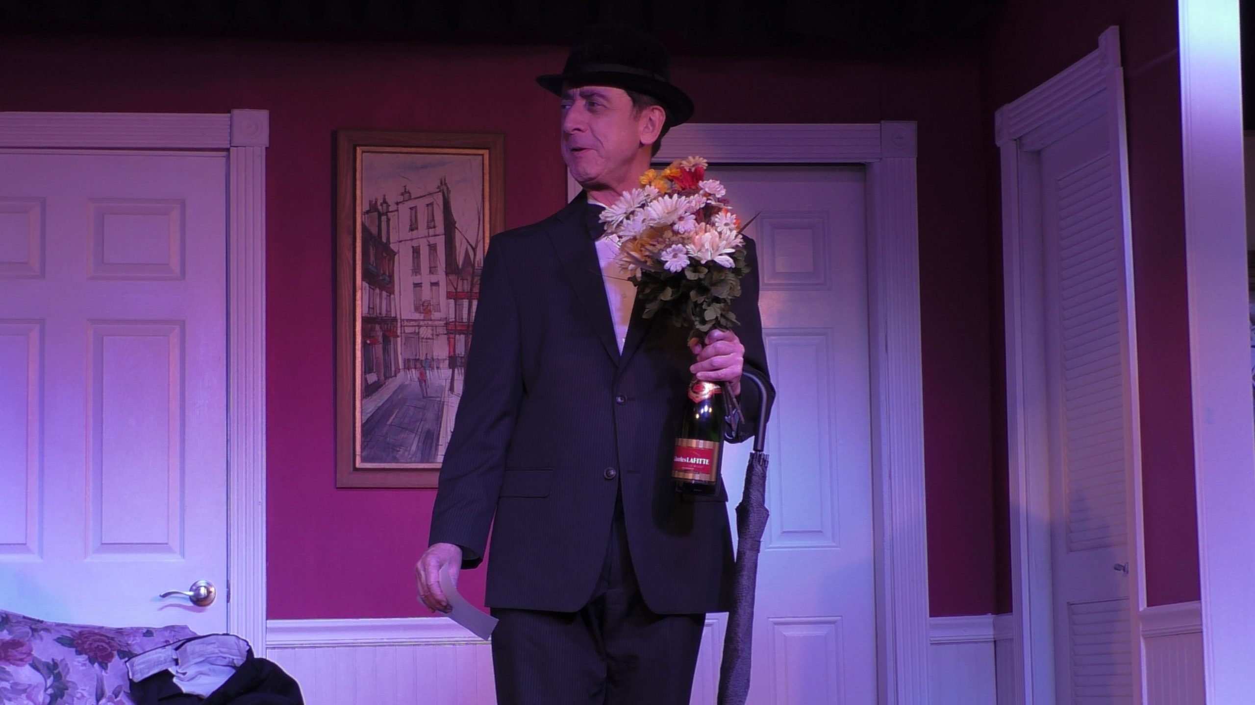 A male actor dressed in a suit and hat holds flowers and a bottle of champagne
