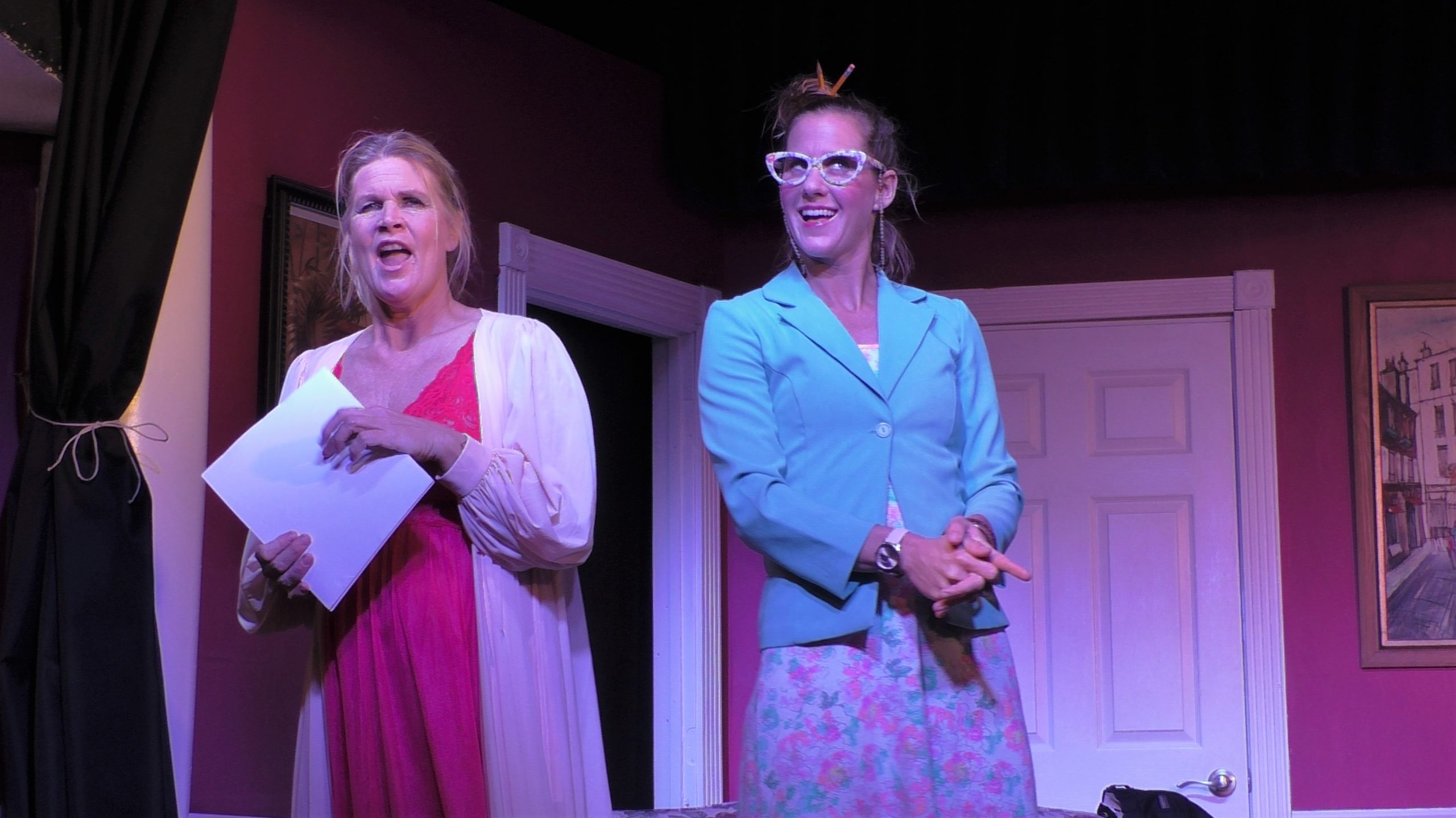 A female actor wears a pink robe and interacts with a female actor who wears a floral skit, green jacket, and large white glasses