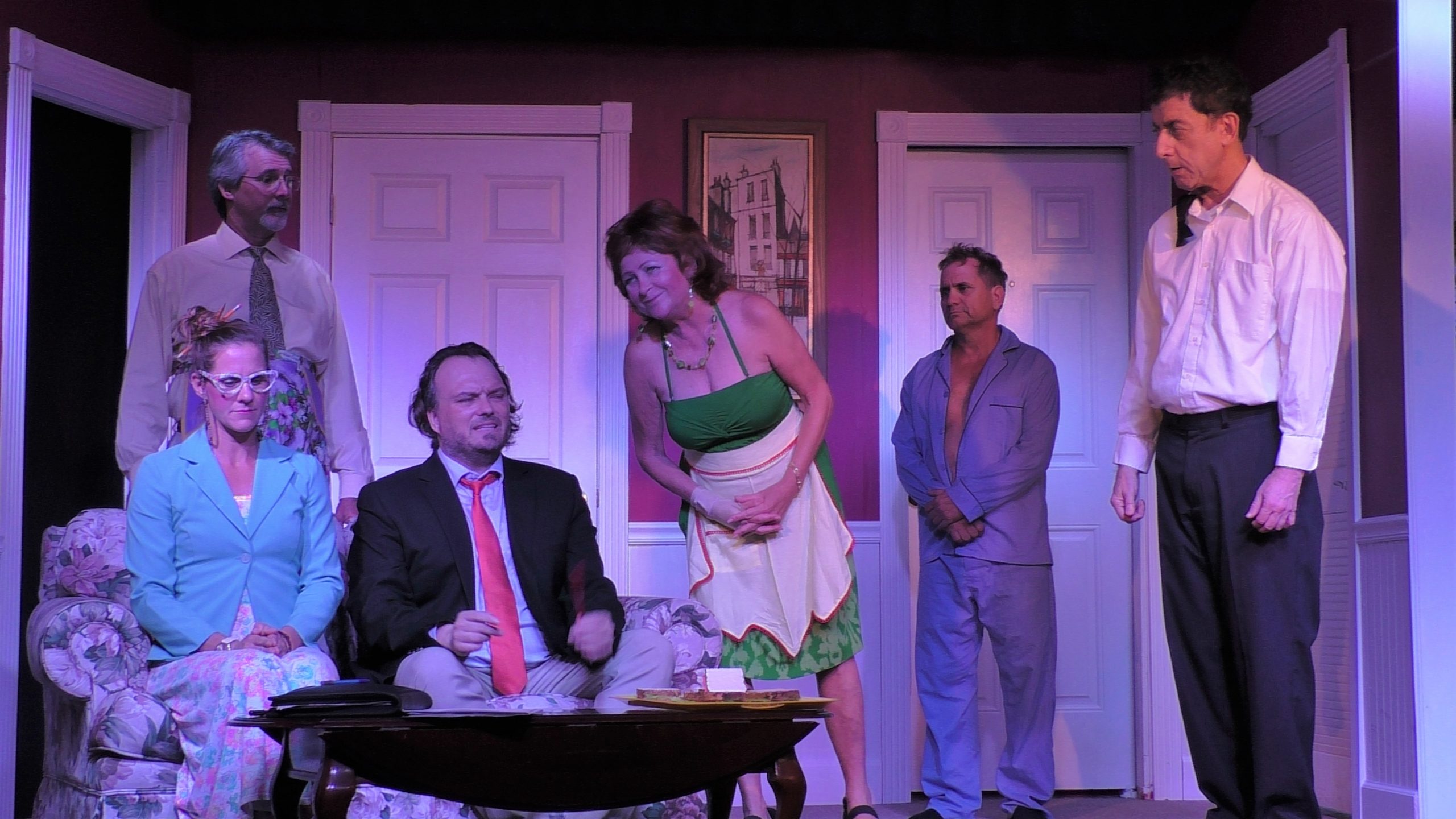 A female wearing a green jack and a male wearing a suite and red tie sit on the sofa and interact with a female wearing a green dress, a male in blue pajamas, and a male in a white shirt and black pants all standing